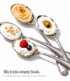 Four silver spoons arranged diagonally on a white background, each holding a creamy food topping: hummus with olive oil and paprika, yogurt with oats, whipped cream with a raspberry and blueberry, and whipped cream with an olive. Text at the bottom reads, 'Mix it into creamy foods.