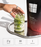 A hand with silver rings holds a glass of green iced protein drink on a silver tray next to a black and red pouch of Pure Base protein isolate. Three white boxes at the bottom display nutritional facts: 27g protein, 110 calories, and 1 ingredient.